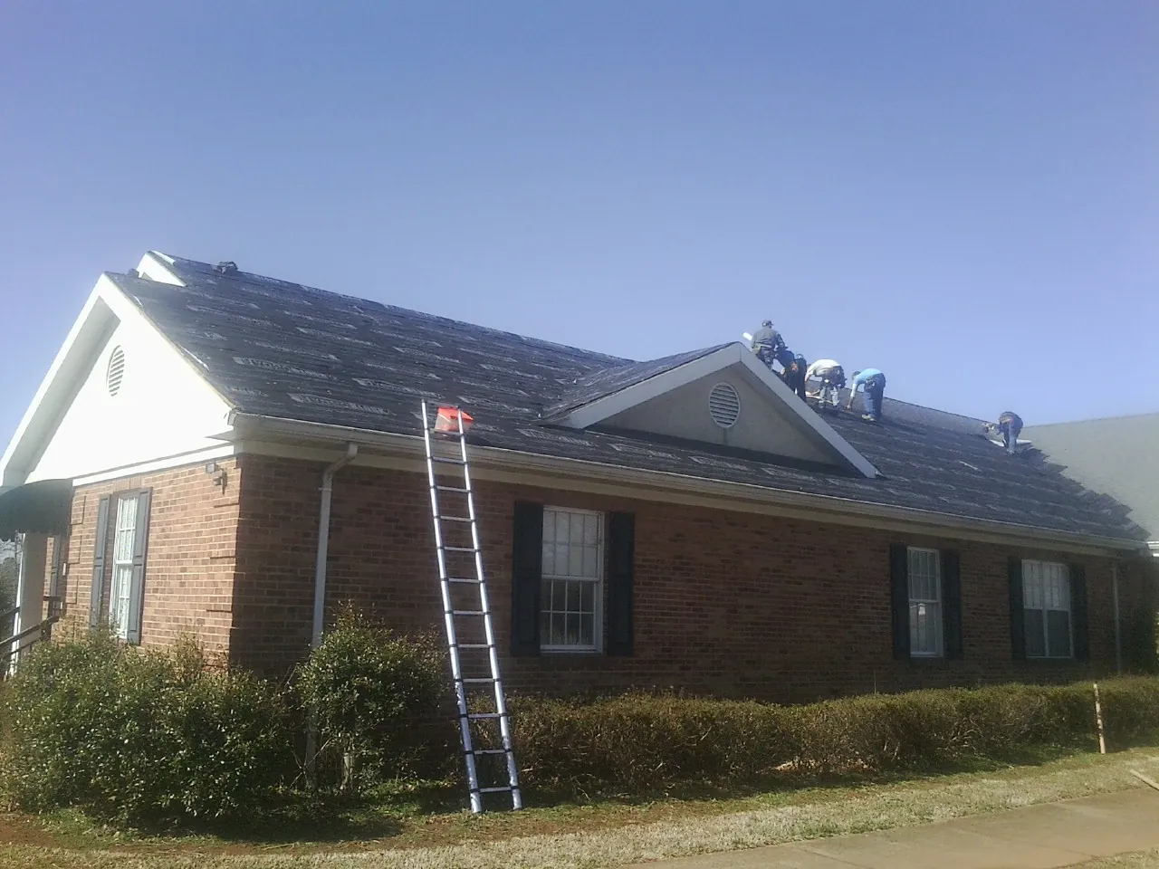 Crew tearing shingles off a brick ranch roof with a ladder set — Metal Roof Repair work by our crew in Overton