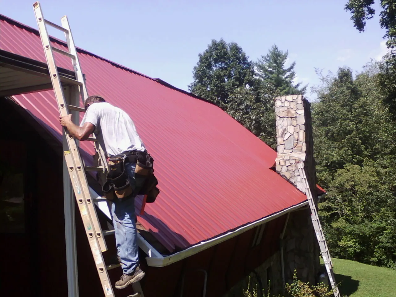 Roofer on a ladder beside a red metal roof and stone chimney — Soffit & Fascia Repair by Roofing Brothers in Overton